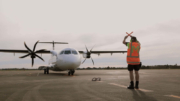 Signal-man wearing high-vis gear standing in front of a newly arrived aeroplane at a Te Utanganui airport.